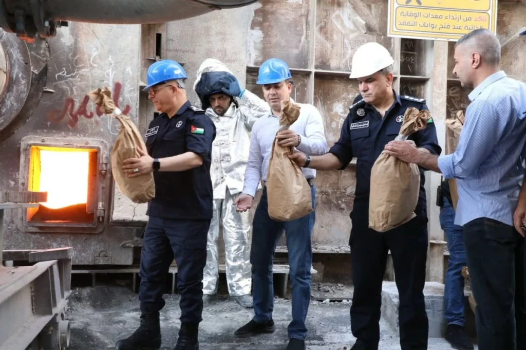 Workers in hard hats and uniforms unloading brown paper bags from a large industrial furnace or kiln. Behind them, a person in a silver protective suit watches.