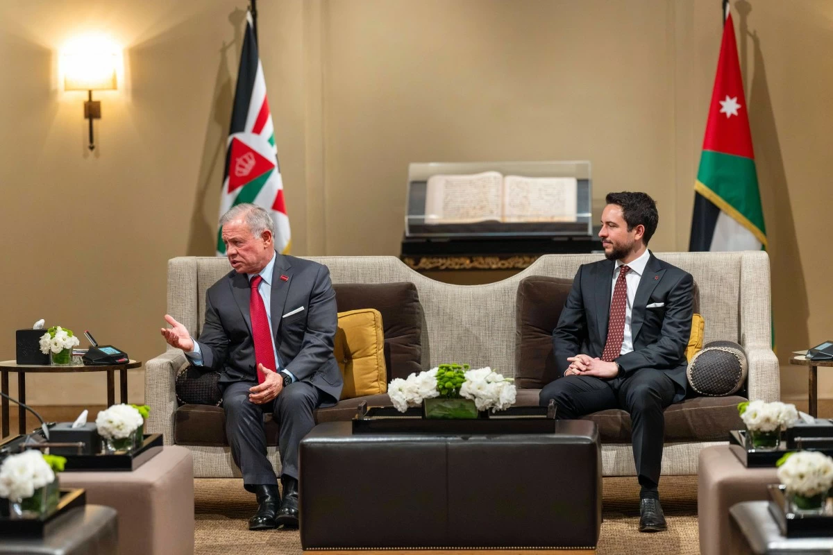 Two men in suits sit on a beige couch in a formal meeting room, with Jordanian flags in the background and a glass-topped table between them.