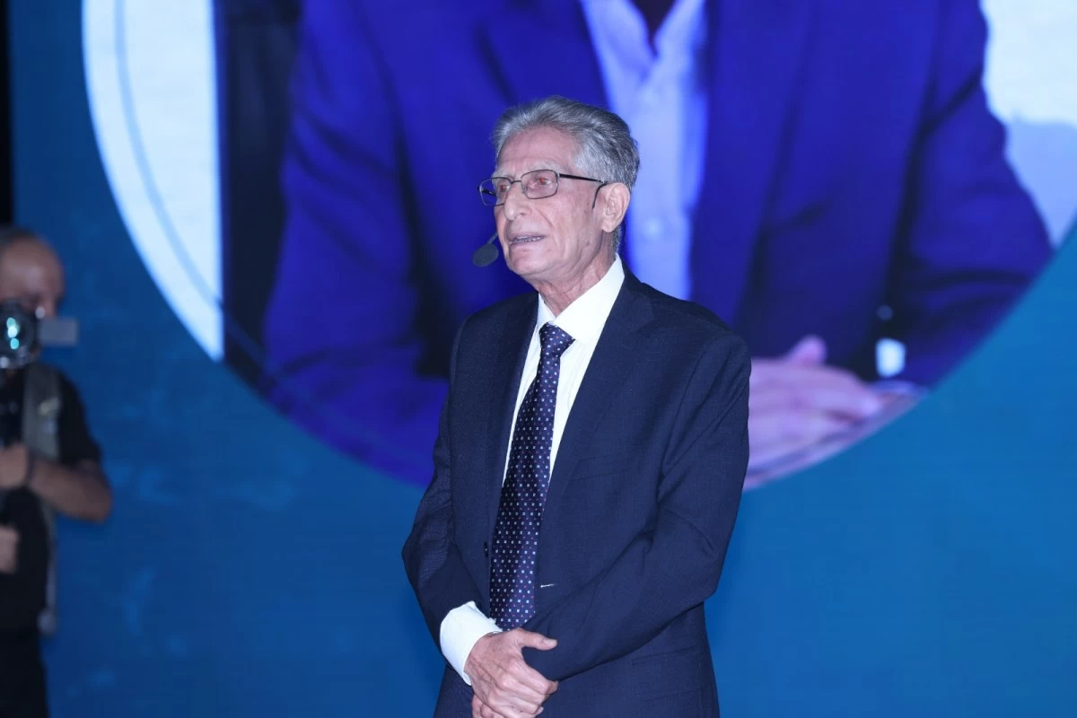 760748 - عروبة الاخباري | Oroba News Older man in a dark suit and polka-dot tie speaks on stage with a headset mic against a blue backdrop.