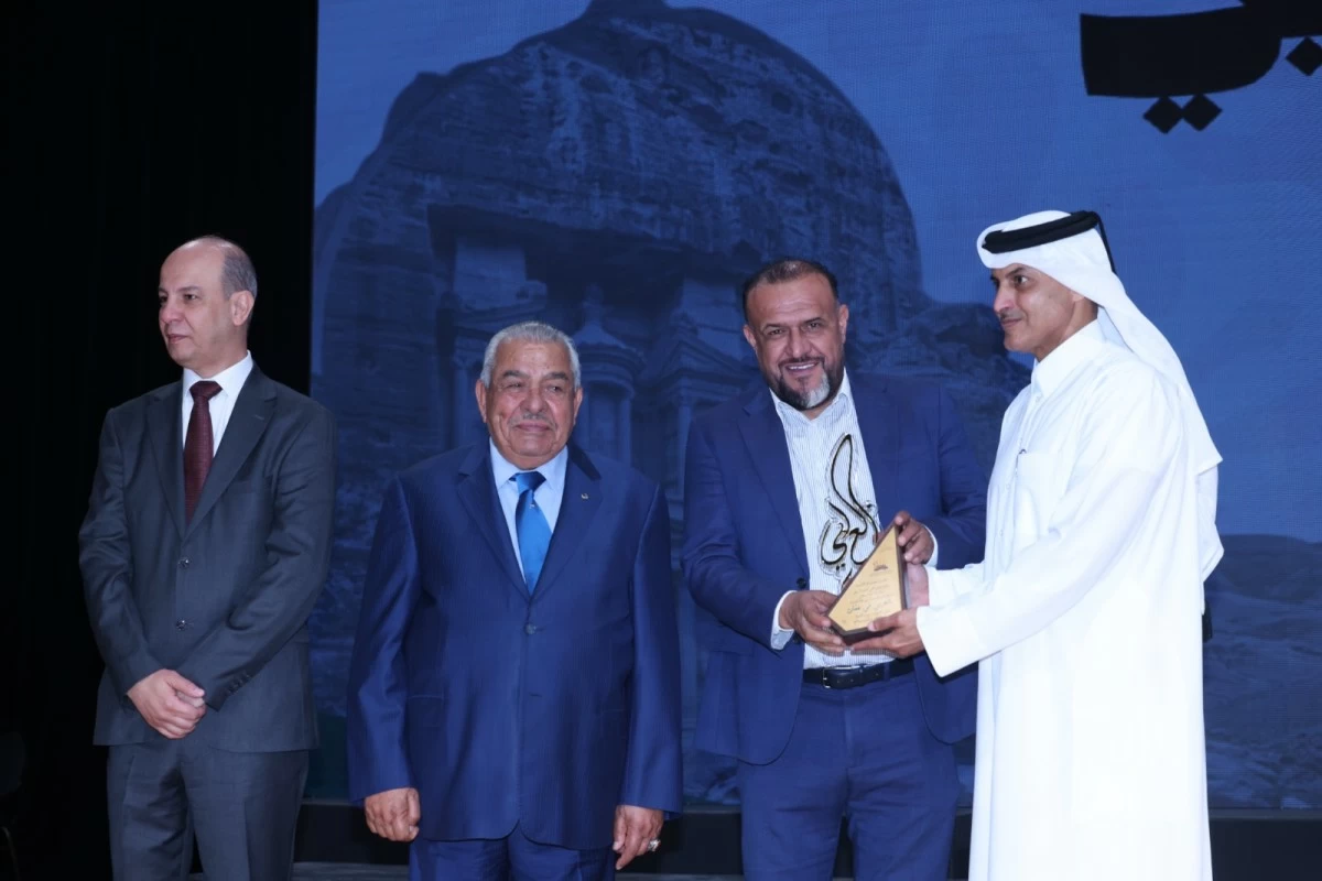 760750 - عروبة الاخباري | Oroba News Four men on a stage; a man in a blue suit receives a triangular trophy from a man in white traditional dress, with two onlookers and a blue backdrop behind them.
