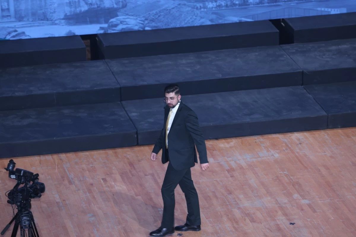 760759 - عروبة الاخباري | Oroba News Man in a dark suit and yellow tie walks across a wooden stage, with a video camera on a tripod nearby and black risers in the background