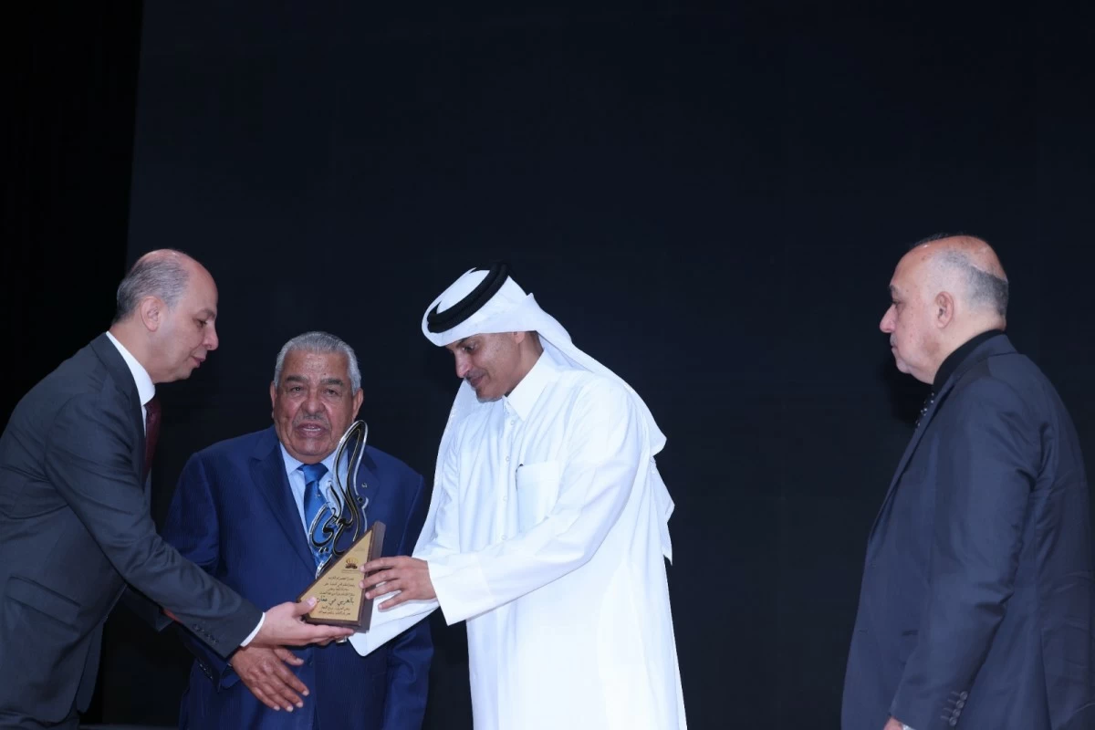 760767 - عروبة الاخباري | Oroba News Man in a blue suit receives an award from a man in traditional white arab dress on a dim stage, with two other men watching on the sides.
