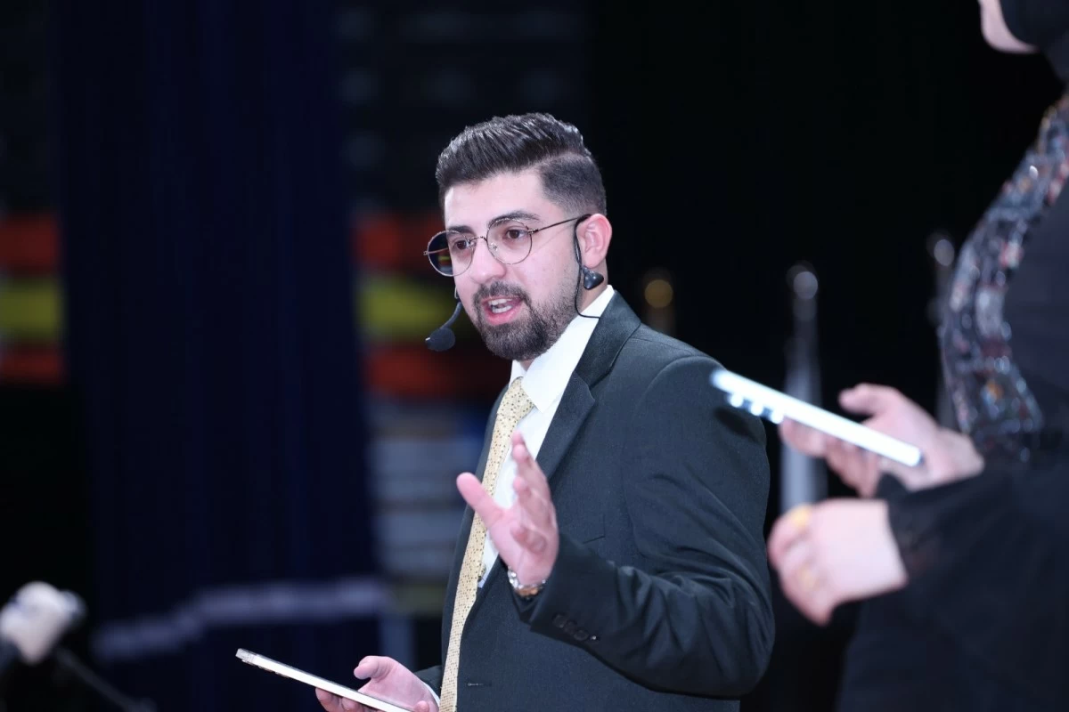 760770 - عروبة الاخباري | Oroba News Man in a dark suit and yellow tie speaks on stage with a headset mic and papers in hand