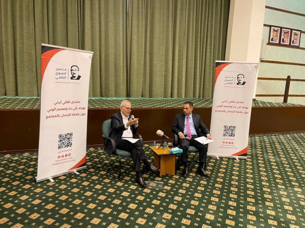 761112 - عروبة الاخباري | Oroba News Two men in suits sit in armchairs on a stage, having a discussion beside a small table with microphones, flanked by tall banners with Arabic text and QR codes on a green carpeted floor.