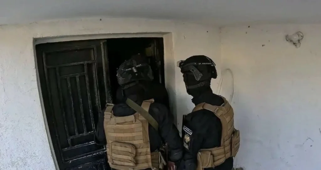 Two armed soldiers in black tactical gear and tan vests approach a dark doorway in a dilapidated building, ready for entry.