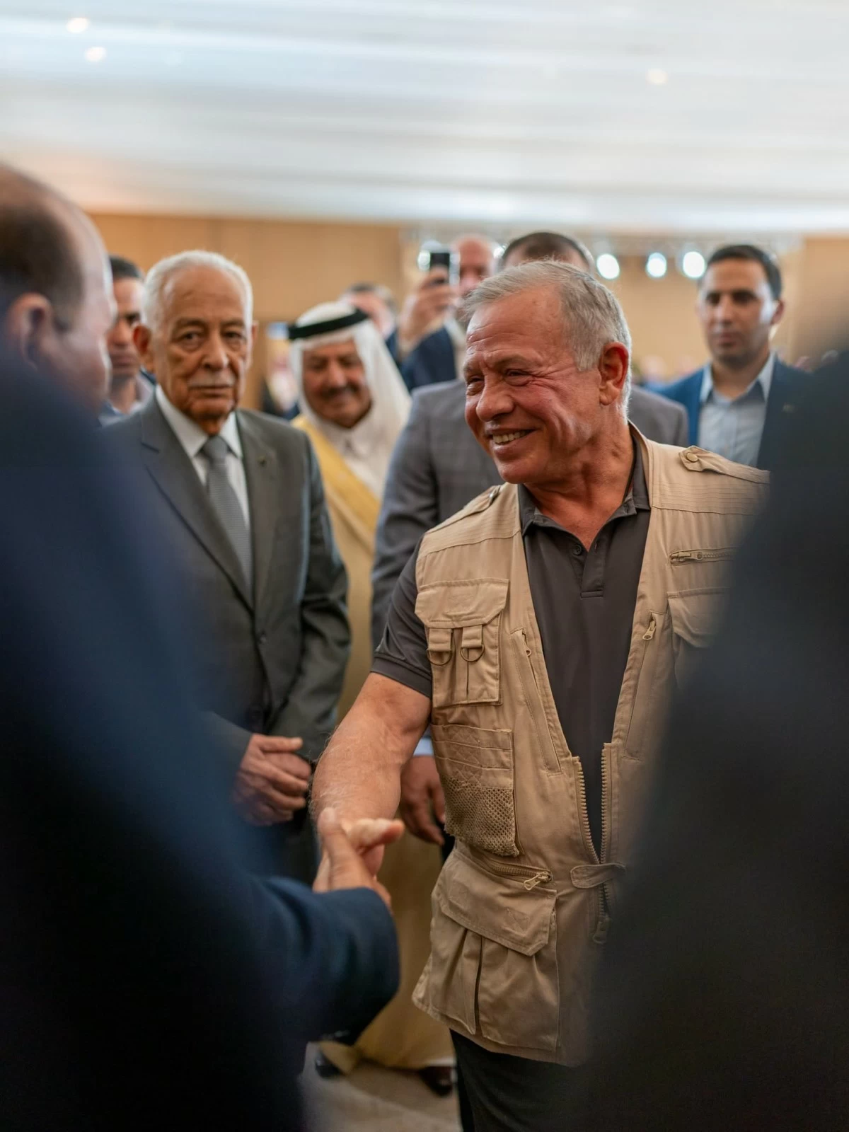 Man in a tan utility vest shaking hands with a suited man at a social or formal event, smiling, with a group of people in the background.