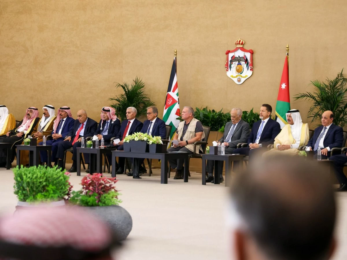 Dignitaries in suits and traditional robes seated in a row at a formal ceremony, with national flags and a crest behind them.