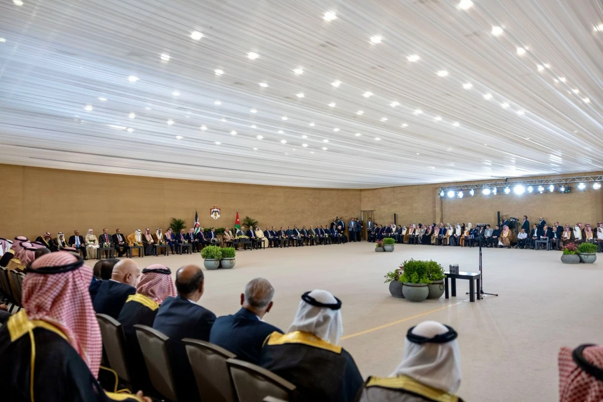 Dignitaries in formal attire sit in a long semicircle inside a large hall, under a white draped ceiling with flags at the back and potted plants in the center.