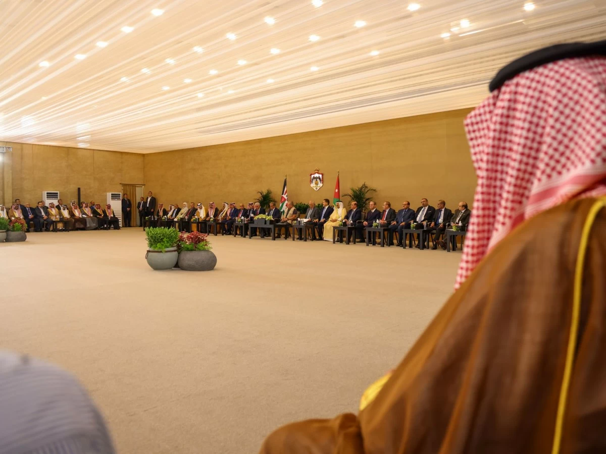Formal meeting in a large hall with officials seated in a long semicircle, flags and an emblem on the wall, and a person in traditional Middle Eastern attire in the foreground.