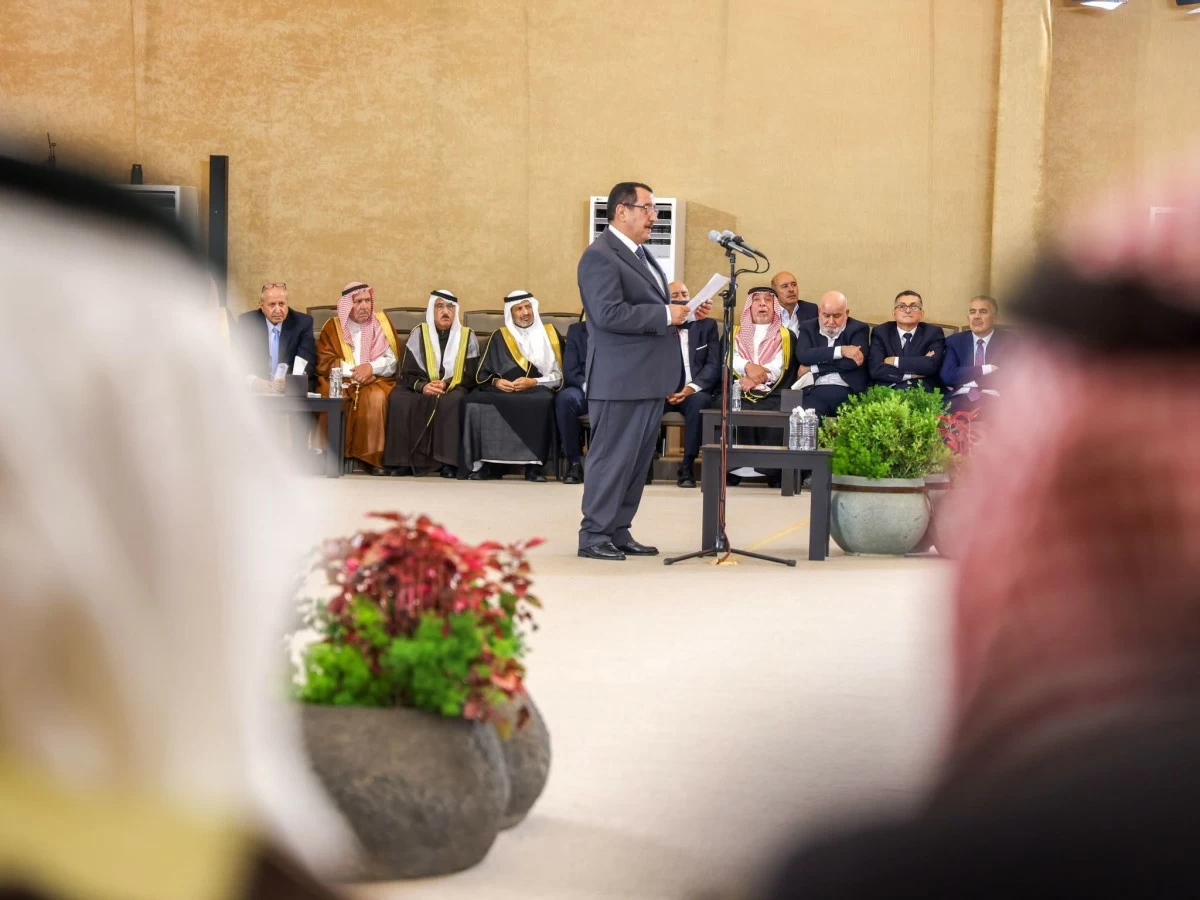 A man in a suit speaks at a microphone stand during a formal ceremony, with dignitaries seated behind him in traditional robes and suits.