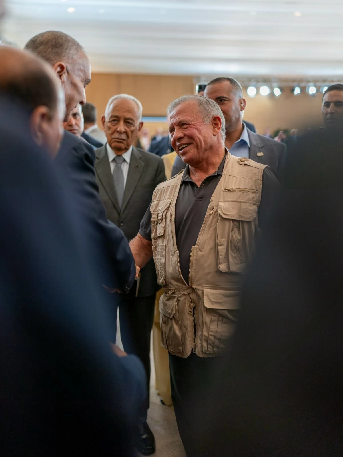 Man in a tan utility vest shakes hands with another man at a crowded indoor event, smiling as others watch.