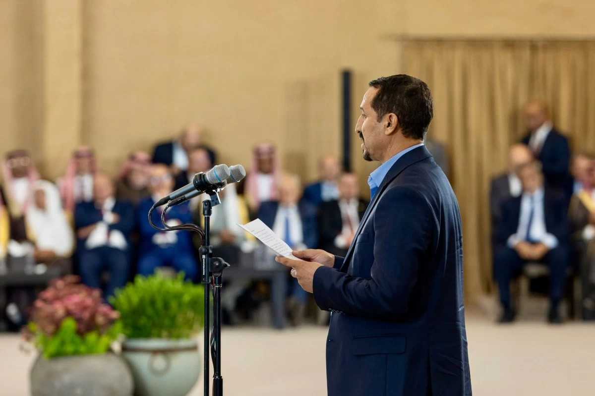 Man in a dark suit delivers a speech at a podium with microphones, holding notes, in a formal event with an audience in the background.