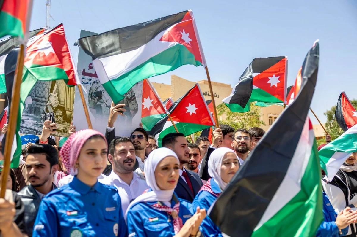 Crowd of protesters waving Jordanian flags on a sunny street; many wear blue uniforms and headscarves.