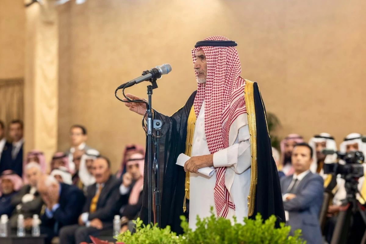 Saudi man in a white thobe and gold-trimmed black bisht speaks into a microphone at a formal ceremony, with an attentive audience behind him.