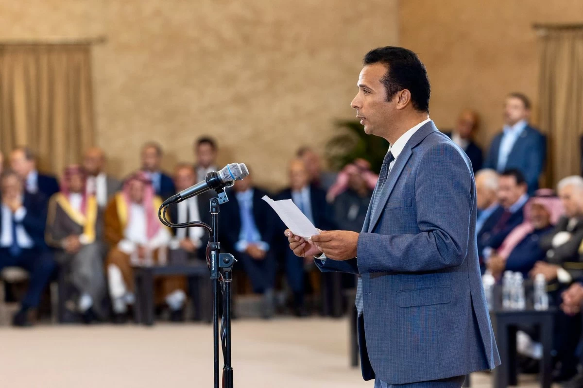 Man in a blue suit delivering a speech at a microphone in a formal event with a seated audience in the background.
