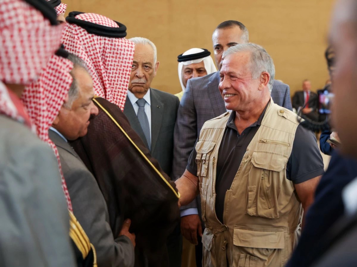 Man in beige utility vest shakes hands with a line of men in traditional Middle Eastern attire at a formal event, smiling broadly.