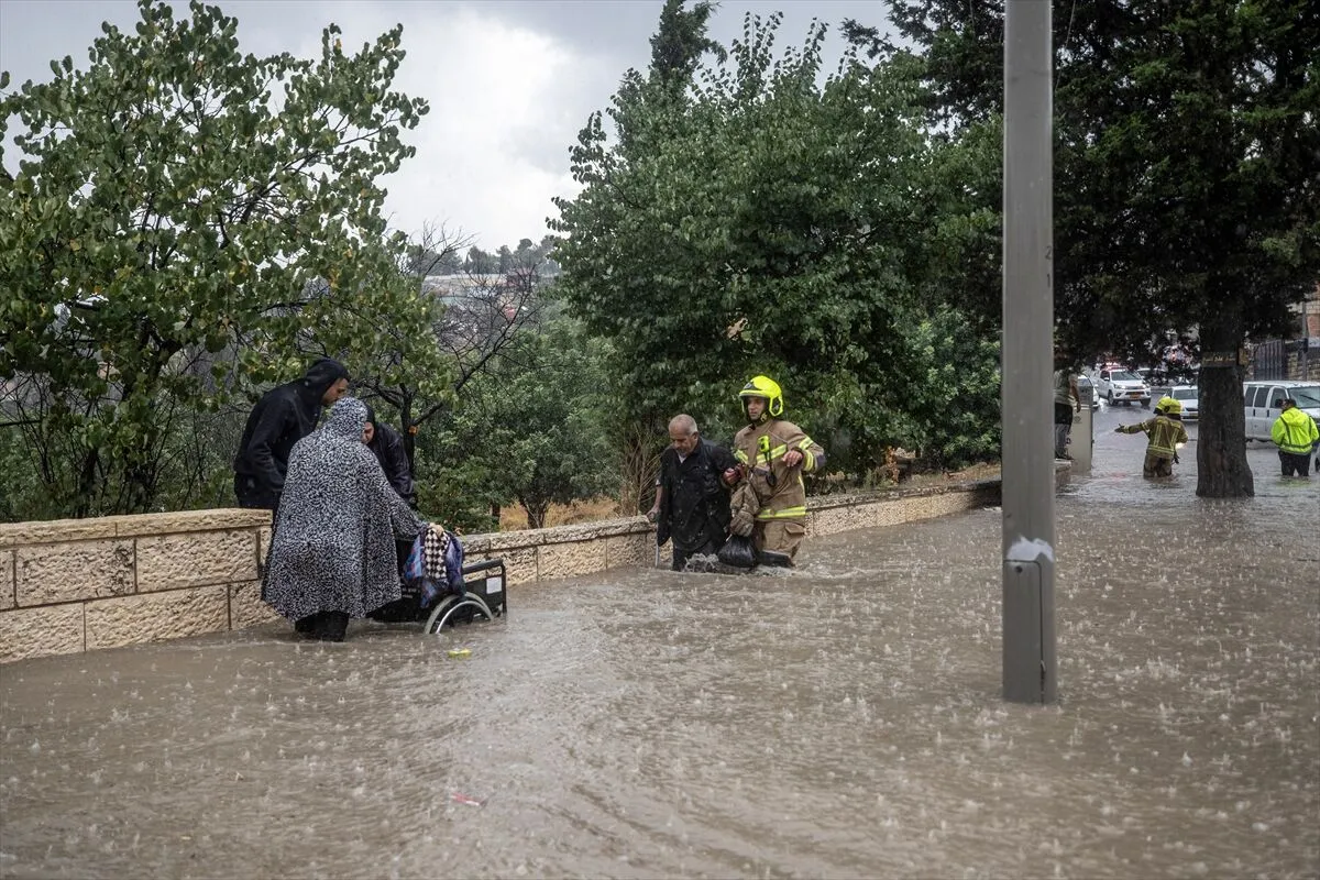 762783 - عروبة الاخباري | Oroba News Rescue workers in yellow gear assist a person in a wheelchair as floodwaters rise on a street; bystanders wade through muddy water.