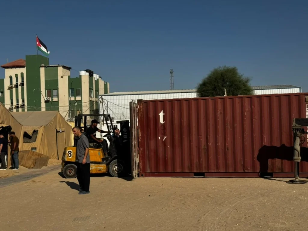 763518 - عروبة الاخباري | Oroba News Desert camp with beige tents, a green building, a forklift, and a red shipping container; Jordanian flag on top.