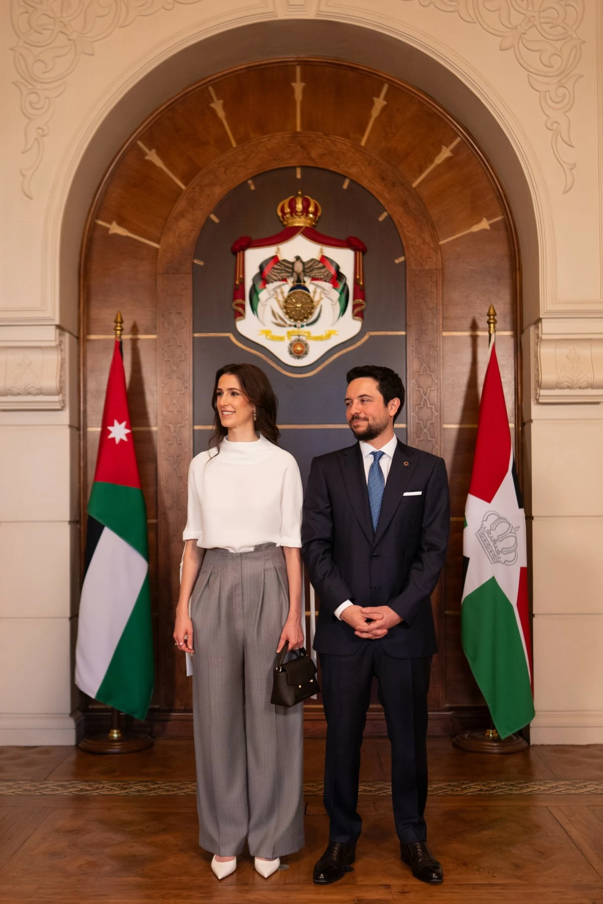 A man in a navy suit and a woman in a white top and gray wide-leg trousers stand side by side in a ceremonial hall with flags and a regal crest behind them.