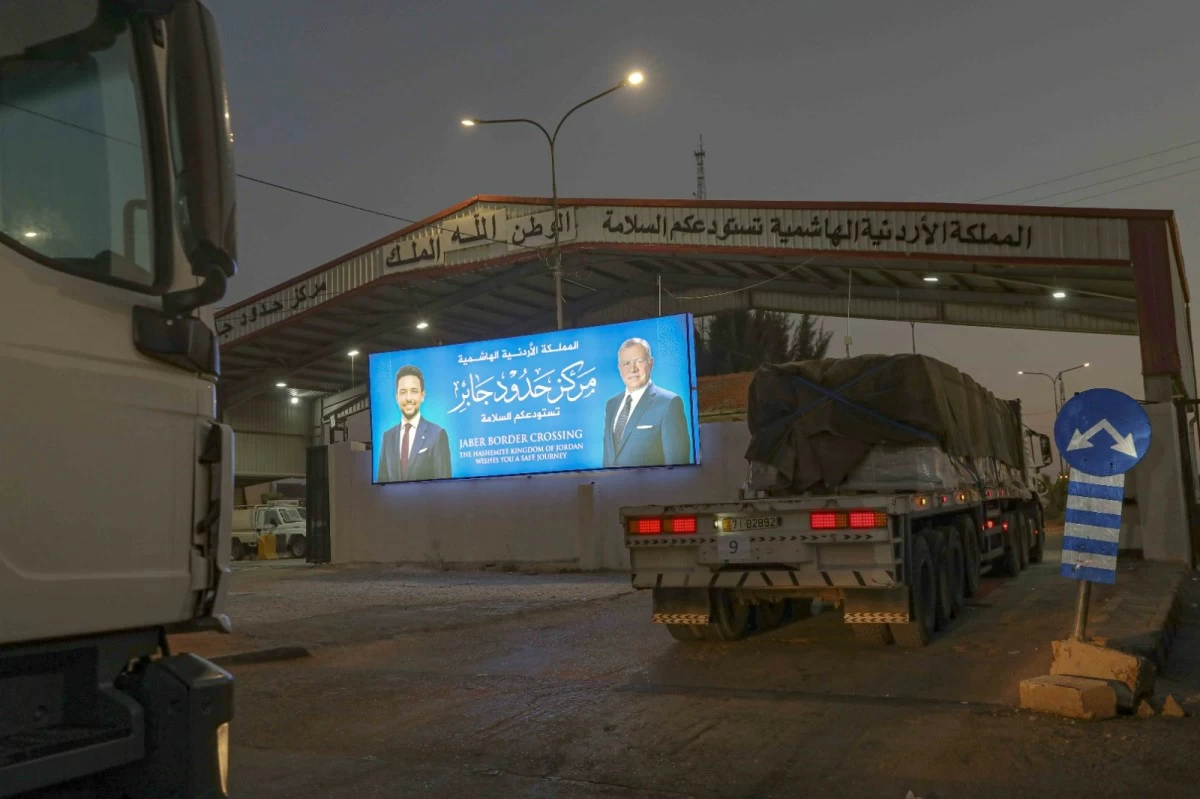 763651 - عروبة الاخباري | Oroba News Border crossing scene at dusk with a large blue billboard showing two men, trucks waiting under Arabic signage.