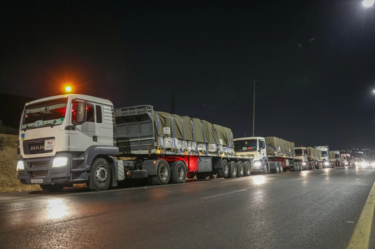 763653 - عروبة الاخباري | Oroba News Line of white heavy-duty trucks with tarped cargo parked on a wet night road, lights reflecting on the pavement.