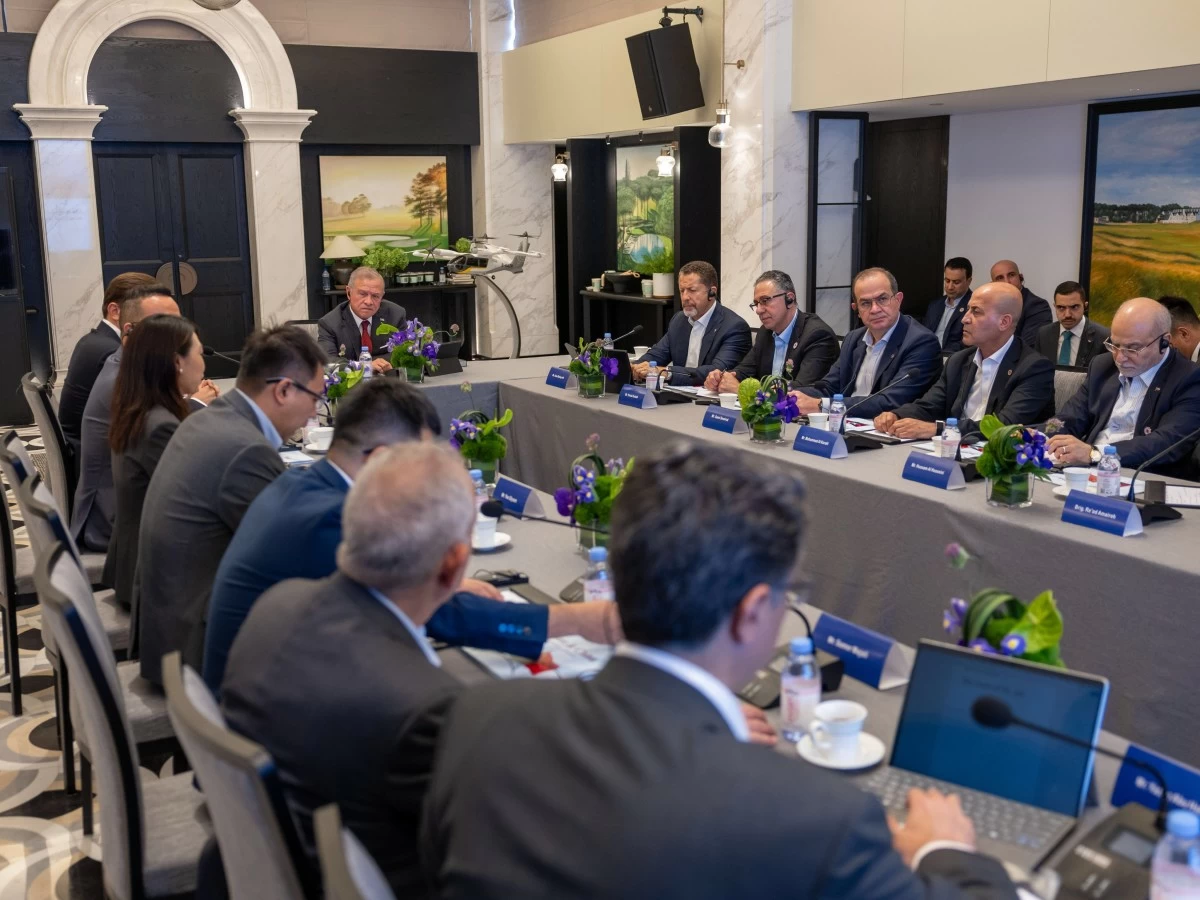 Business meeting with multiple executives seated around a long table, laptops open, nameplates, and purple flower centerpieces in a formal conference room.
