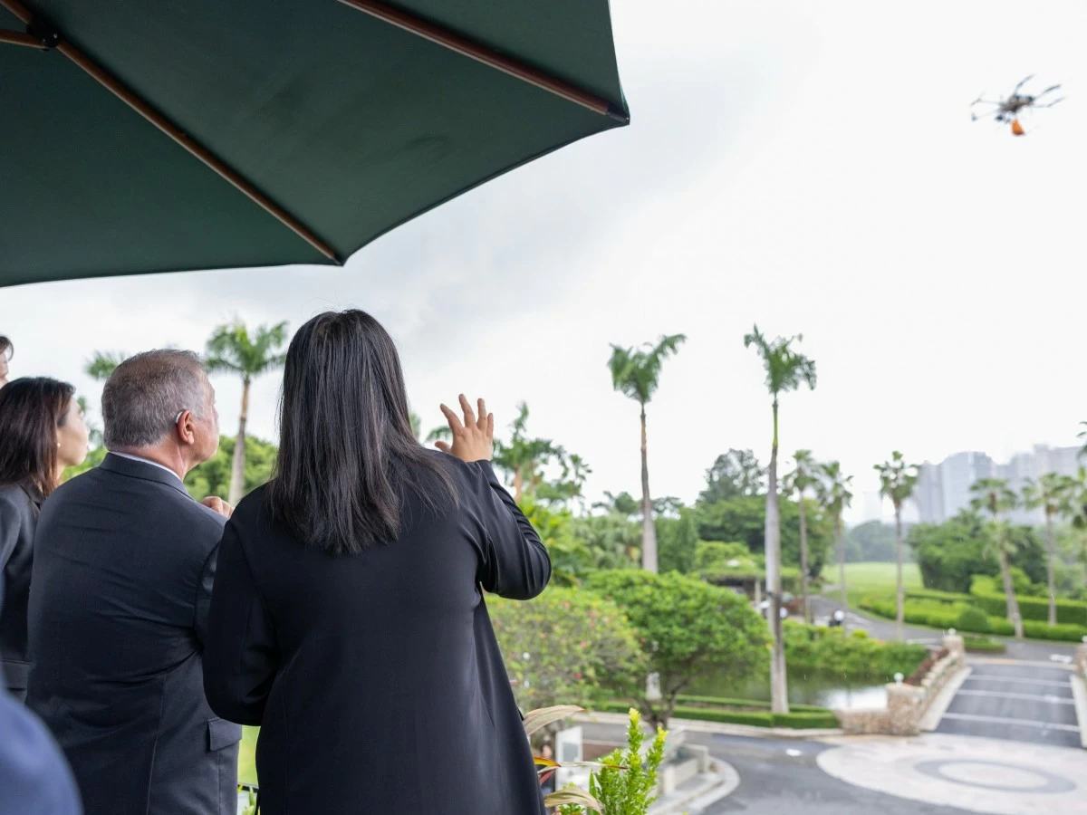 Group of professionals in suits on a terrace, watching a drone fly overhead among palm trees.