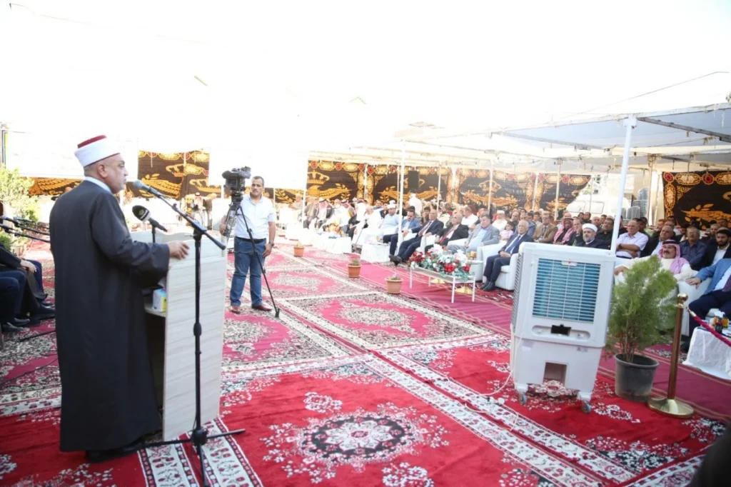 Religious leader in a robe and white hat speaks from a podium to a seated audience under a tent.