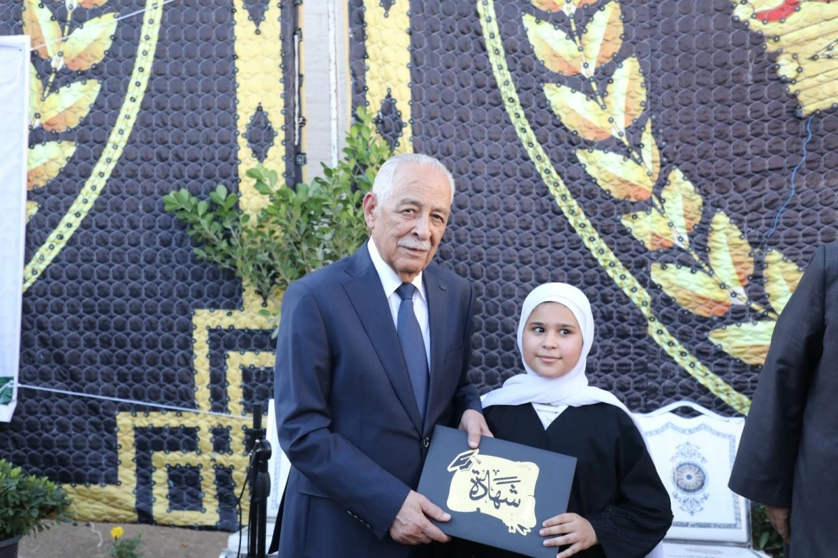 Older man in a suit presents a certificate to a girl in a white hijab at an outdoor ceremony with a decorative backdrop behind them.