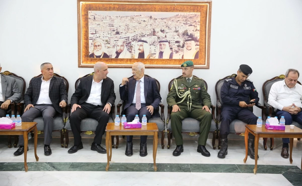 Group of officials in suits and military uniforms seated along a row in a meeting room, with a framed photo on the wall behind them.