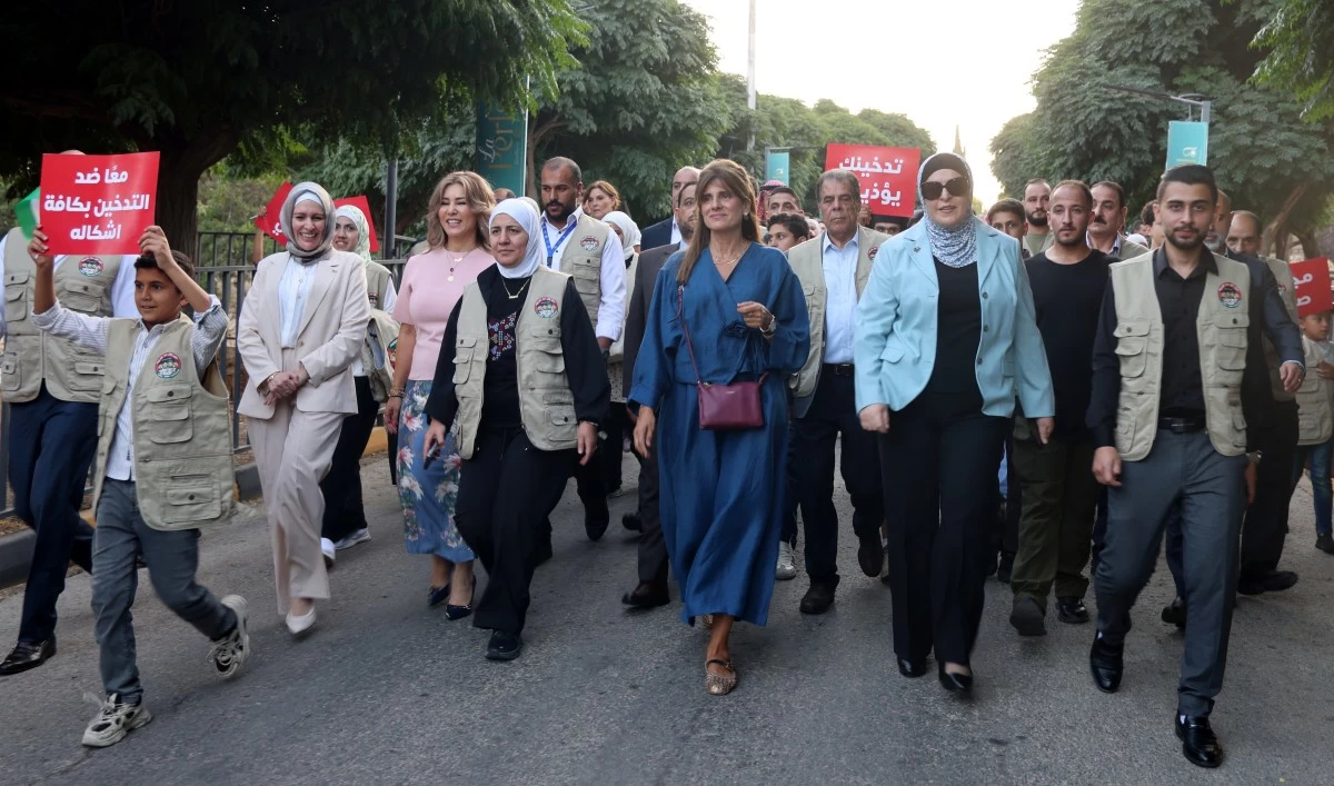 Group of diverse protesters walking down a street, many wearing beige vests, with red Arabic signs in the background.