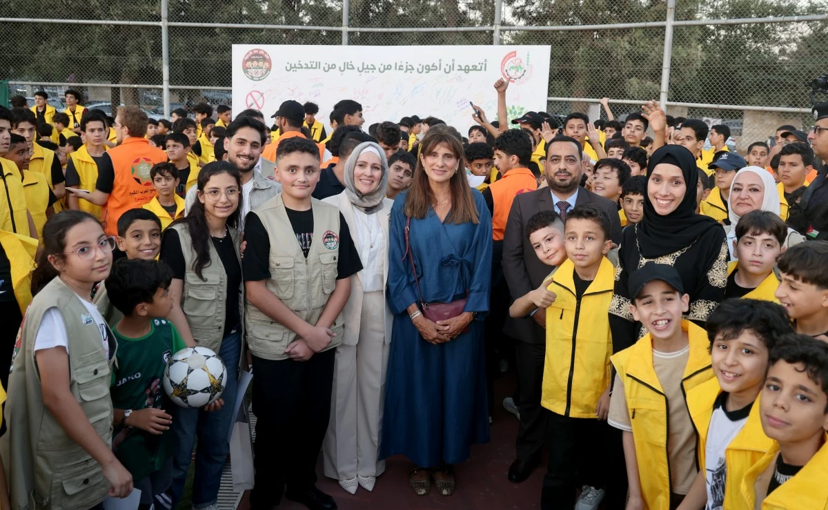Group of smiling kids and adults posing for a photo at an outdoor event, many in yellow vests; a banner with Arabic text behind them.