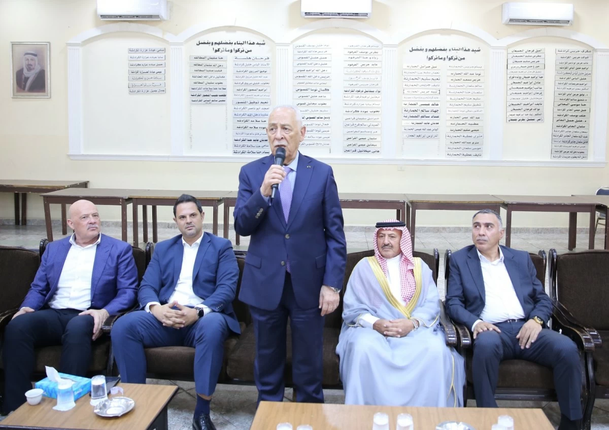 764760 - عروبة الاخباري | Oroba News A man in a blue suit speaks into a microphone while four men sit in a row in a formal meeting room, with Arabic posters on the wall behind them.