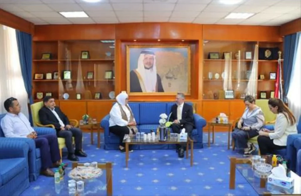 Six people in formal attire sit on blue sofas in a meeting room, with a large framed portrait on the wall behind them.