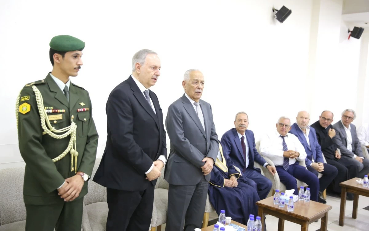 765043 - عروبة الاخباري | Oroba News Uniformed officer in green stands beside three men in suits at a formal meeting, with other officials seated in a row behind them in a conference room.