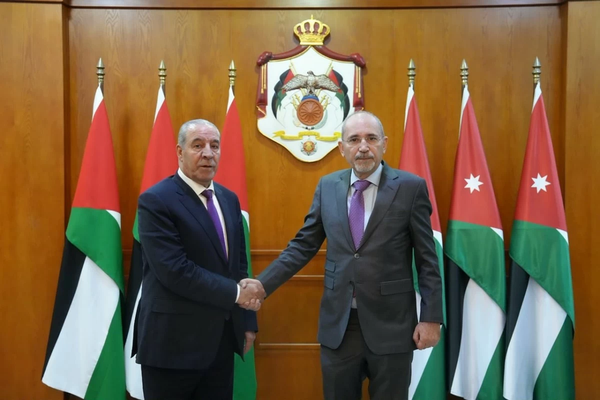 Two men in business suits shake hands in a formal room, with Jordanian flags and a national crest behind them.
