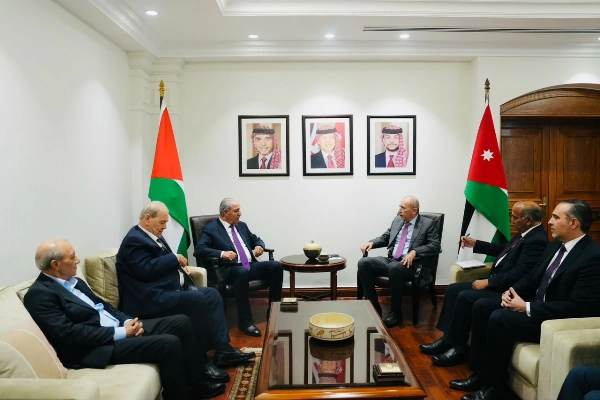 Six men in business suits sit in a formal meeting room with Jordanian flags on either side and three framed portraits on the wall above a central coffee table.