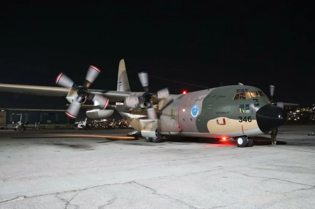 Nighttime airfield scene with a four-engine propeller military transport aircraft on the tarmac, red beacon lights visible by the nose gear.