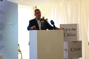 Man in a blazer speaks at a podium with multiple microphones, white curtains in the background and branded Qatrana Cement boxes beside him.
