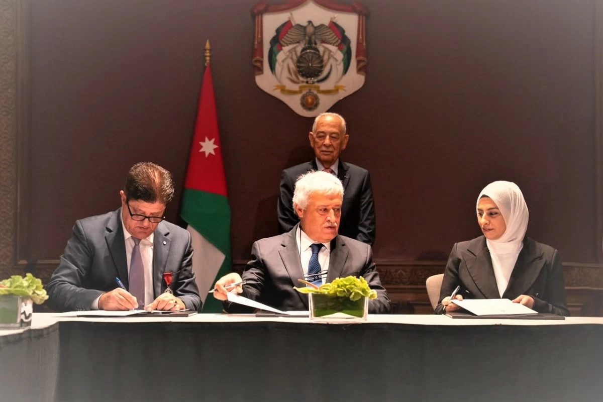 765699 - عروبة الاخباري | Oroba News Three officials sign documents at a formal ceremony; Jordan flag and national emblem behind them, central figure signing.