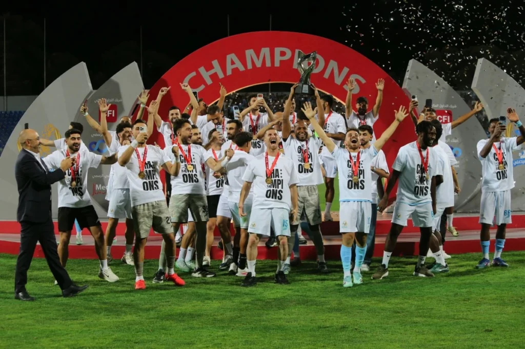 765910 - عروبة الاخباري | Oroba News Soccer team celebrating on the field after victory, lifting the trophy with medals around their necks under a red champions arch behind them, confetti in the air.