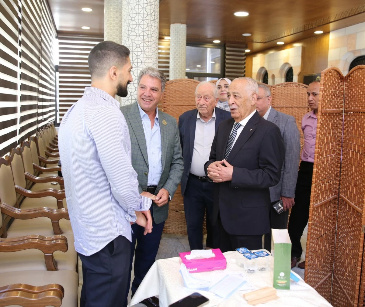 766041 - عروبة الاخباري | Oroba News Group of men in suits chat with a younger man in a striped shirt in a lobby with wicker partitions and row seating nearby.