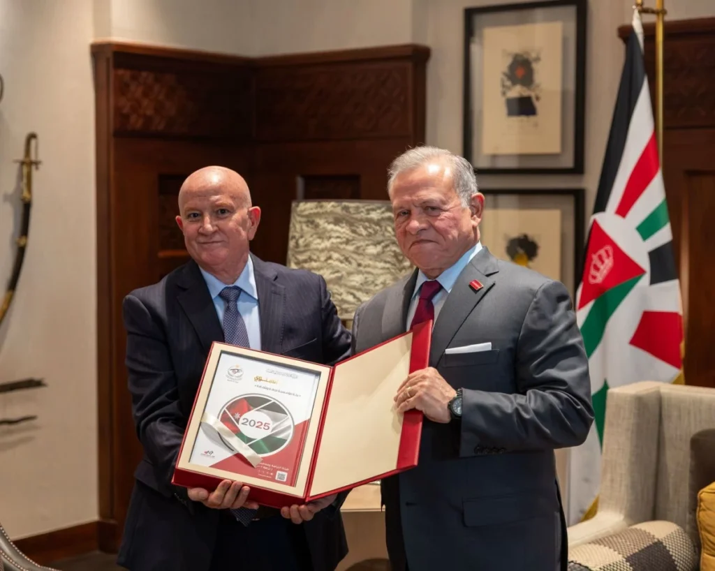 766268 - عروبة الاخباري | Oroba News Two men in suits hold open red folder presenting a framed certificate during a formal award ceremony, flag in background.