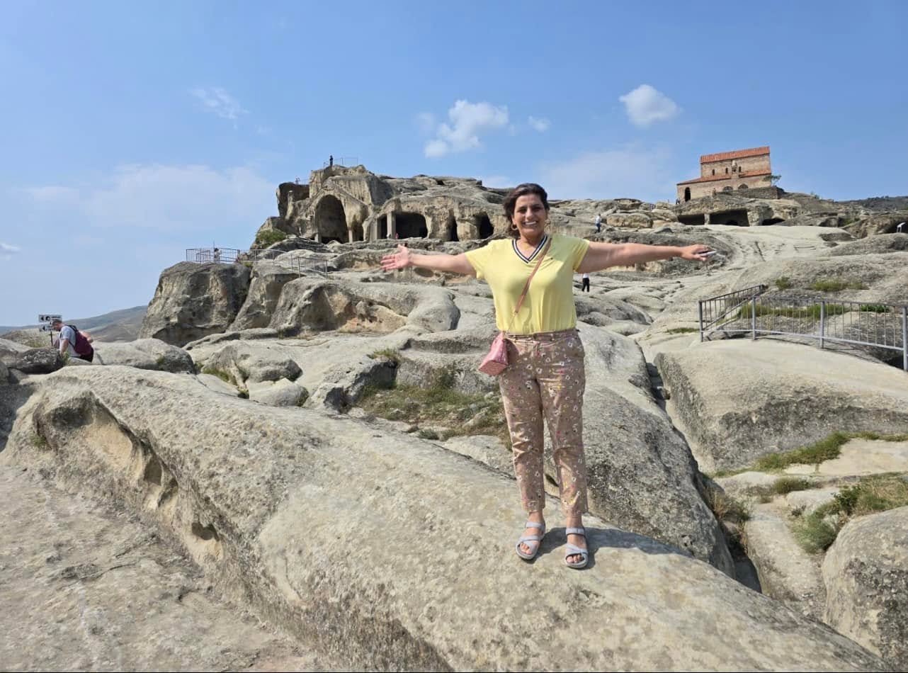 Screenshot - عروبة الاخباري | Oroba News Smiling woman in a yellow top and patterned pants stands on large rocks with arms outstretched; ancient cave dwellings and a brick ruin loom in the background under a blue sky.