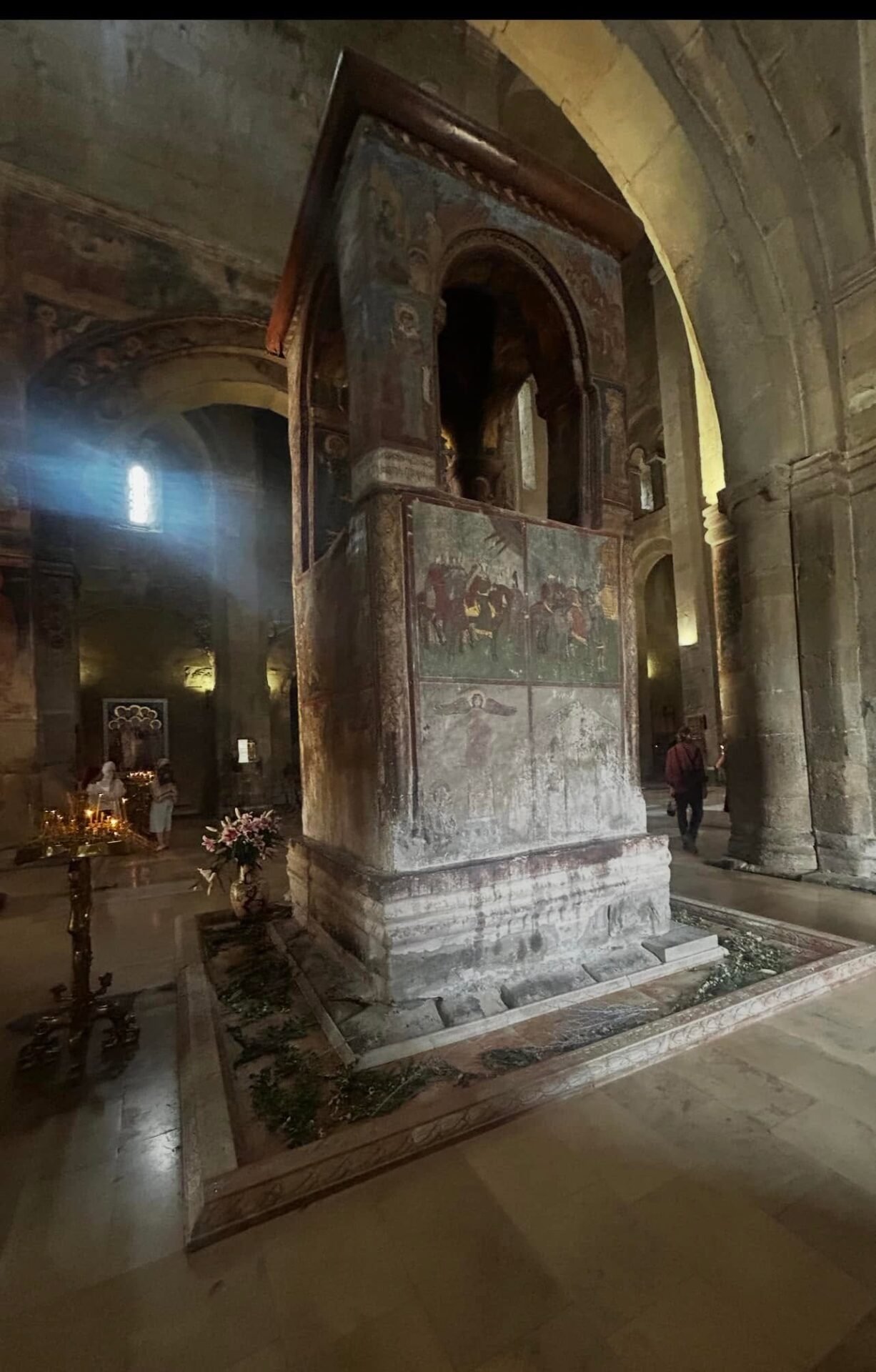 Screenshot - عروبة الاخباري | Oroba News Interior of an old stone church with a tall painted column and arches, sunlight streaming through a narrow window onto the frescoed surfaces and floor.