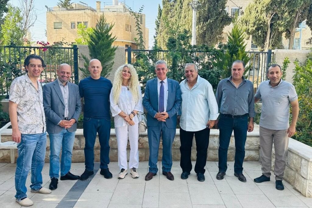 767031674_10243781913873446_7164150434908180388_n 1 - عروبة الاخباري | Oroba News Eight adults stand in a paved courtyard posing for a group photo, with casual and business attire and greenery in the background.