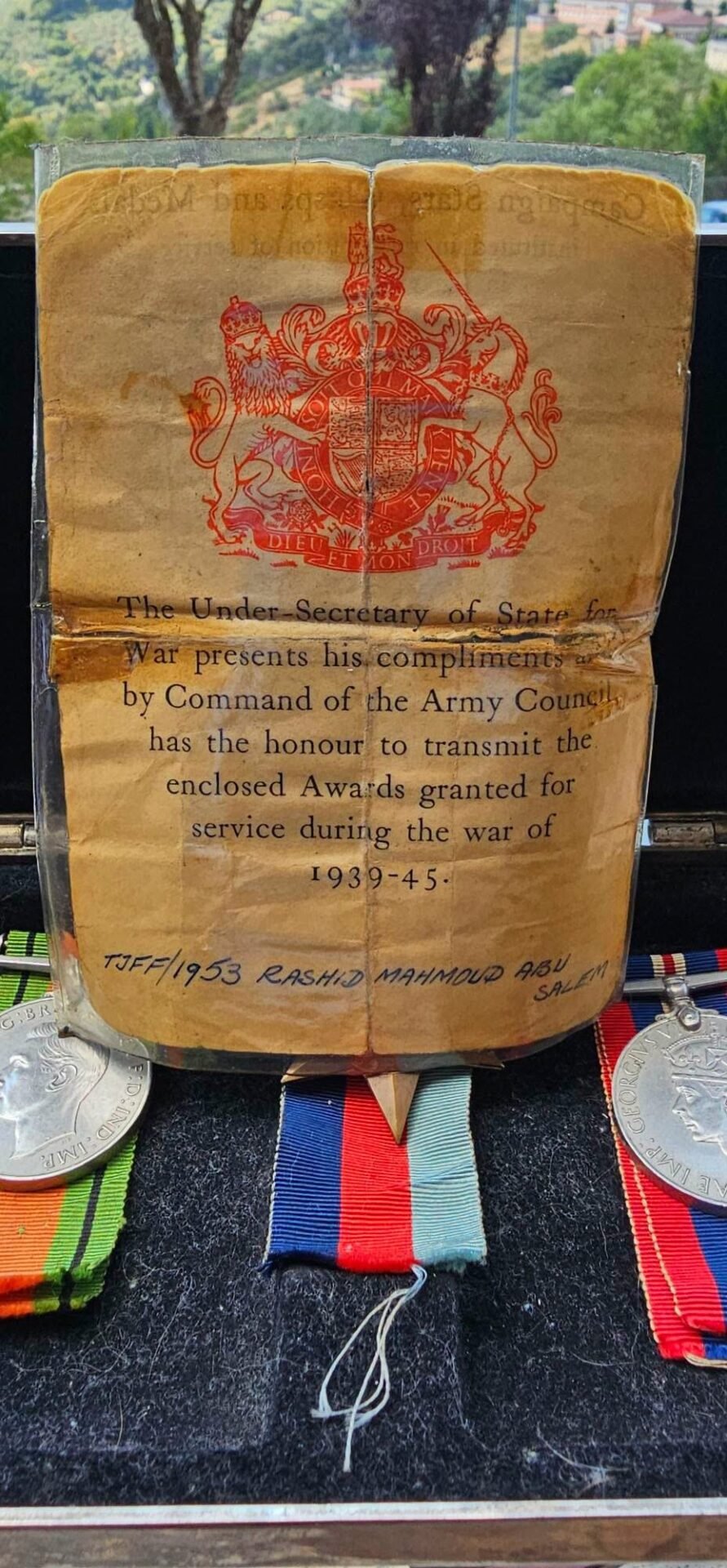 Worn old certificate with a red royal crest and the lines about awards for service during the war (1939-45), laid over a display with ribbons and medals below.