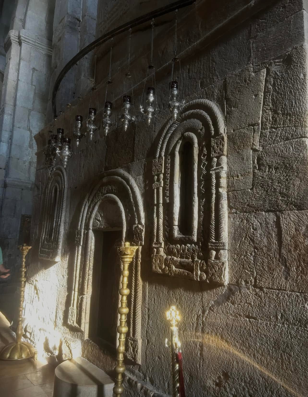 Screenshot - عروبة الاخباري | Oroba News Stone church wall with carved arched niches and small window, lined by hanging oil lamps and brass candleholders along a stone altar area.”,