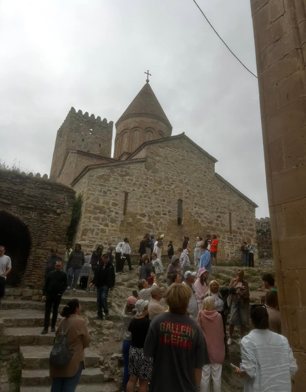 Screenshot - عروبة الاخباري | Oroba News Stone church with a conical roof and cross, set in a stone-walled historic site with a crowd of visitors outside under a cloudy sky.