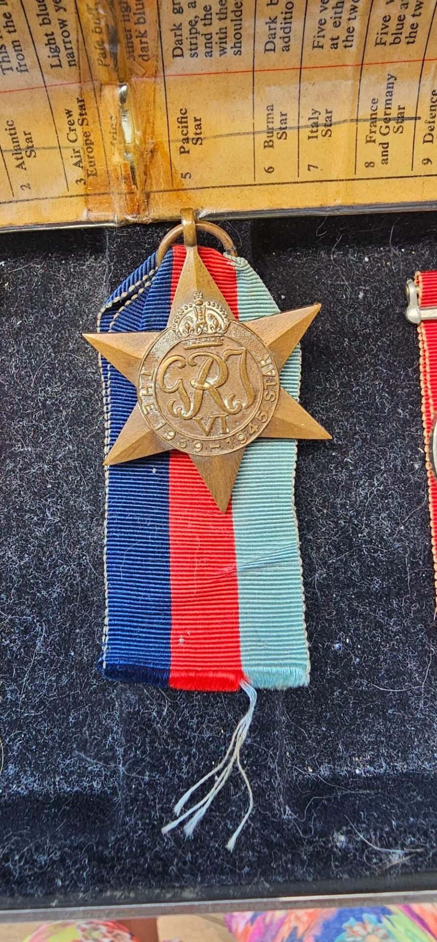 Bronze five-point star medal with a crowned circular center and monogram, suspended from a blue–red–light blue striped ribbon on a dark fabric backdrop.